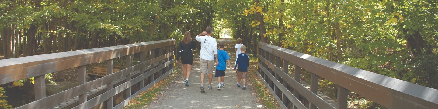 Family walks across a bridge over the Clinton River into a wooded path